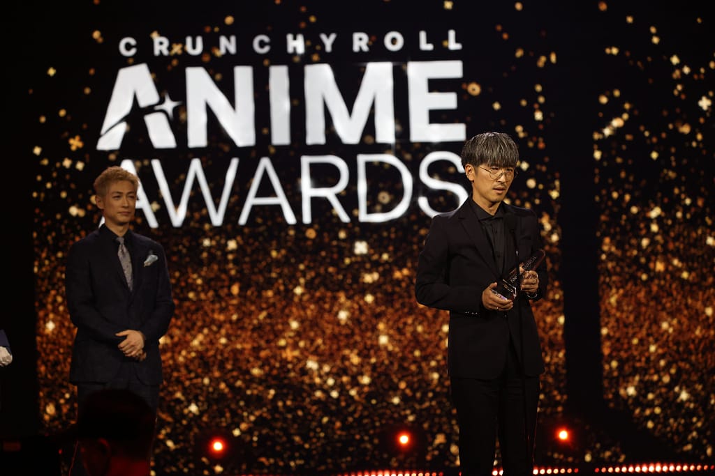 Un hombre con gafas sostiene un premio en el escenario de los Crunchyroll Anime Awards, mientras otro hombre observa al fondo. El fondo está decorado con luces brillantes.