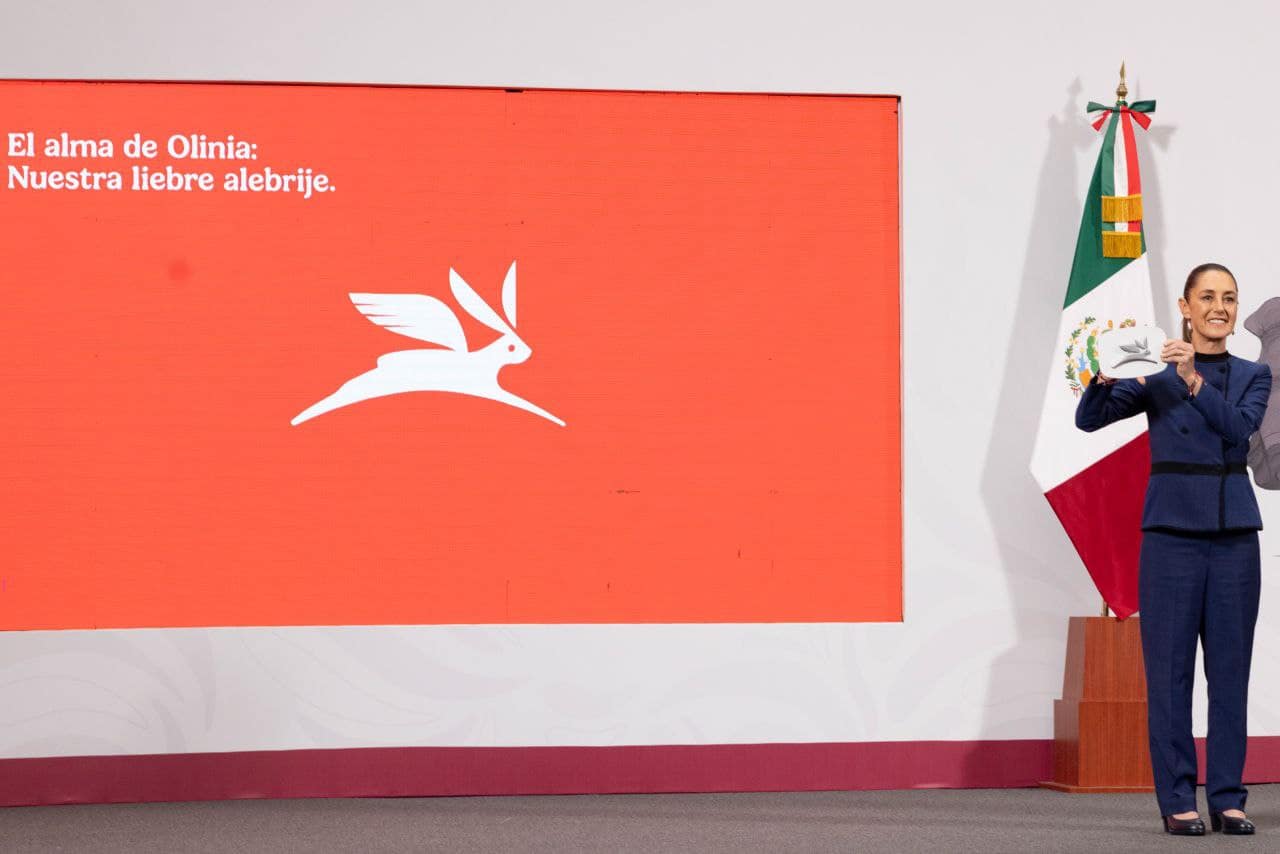 Mujer en un evento sosteniendo un objeto con un diseño de un alebrije, con un fondo naranja y texto que dice 'El alma de Olinia: Nuestra libre alebrije'. También se puede ver una bandera de México al fondo.