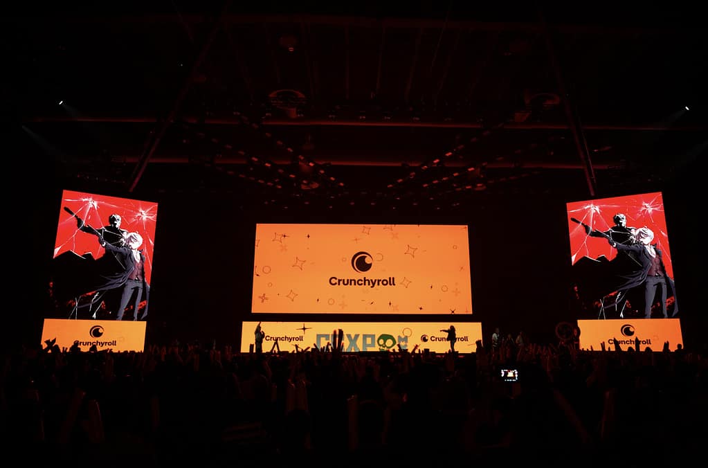 Escenario de un evento de Crunchyroll con grandes pantallas mostrando gráficos animados. La audiencia está animada, con muchas manos levantadas.