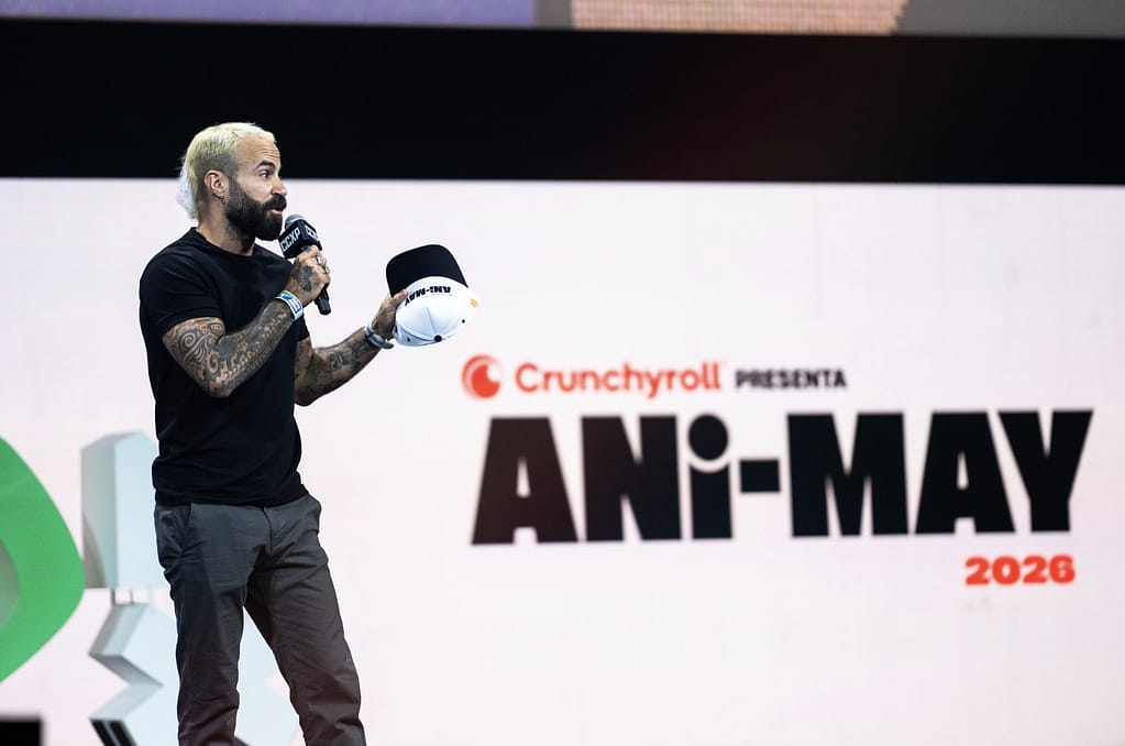 Un presentador sosteniendo un micrófono y una gorra en el escenario, con un fondo que muestra el logo de 'Ani-May 2026' y el nombre de Crunchyroll.