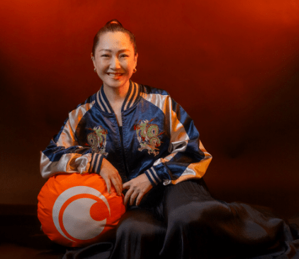 Mujer sonriente sentada junto a un cojín naranja con el logo de Crunchyroll, vestida con una chaqueta brillante de colores en un fondo oscuro.